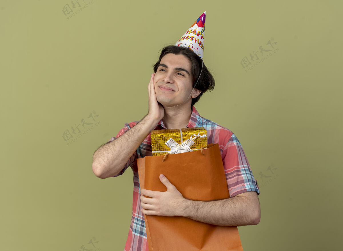 绿色戴着生日帽的白人帅哥高兴地把手放在脸上 手里拿着购物袋里的礼品盒盒子穿拿着