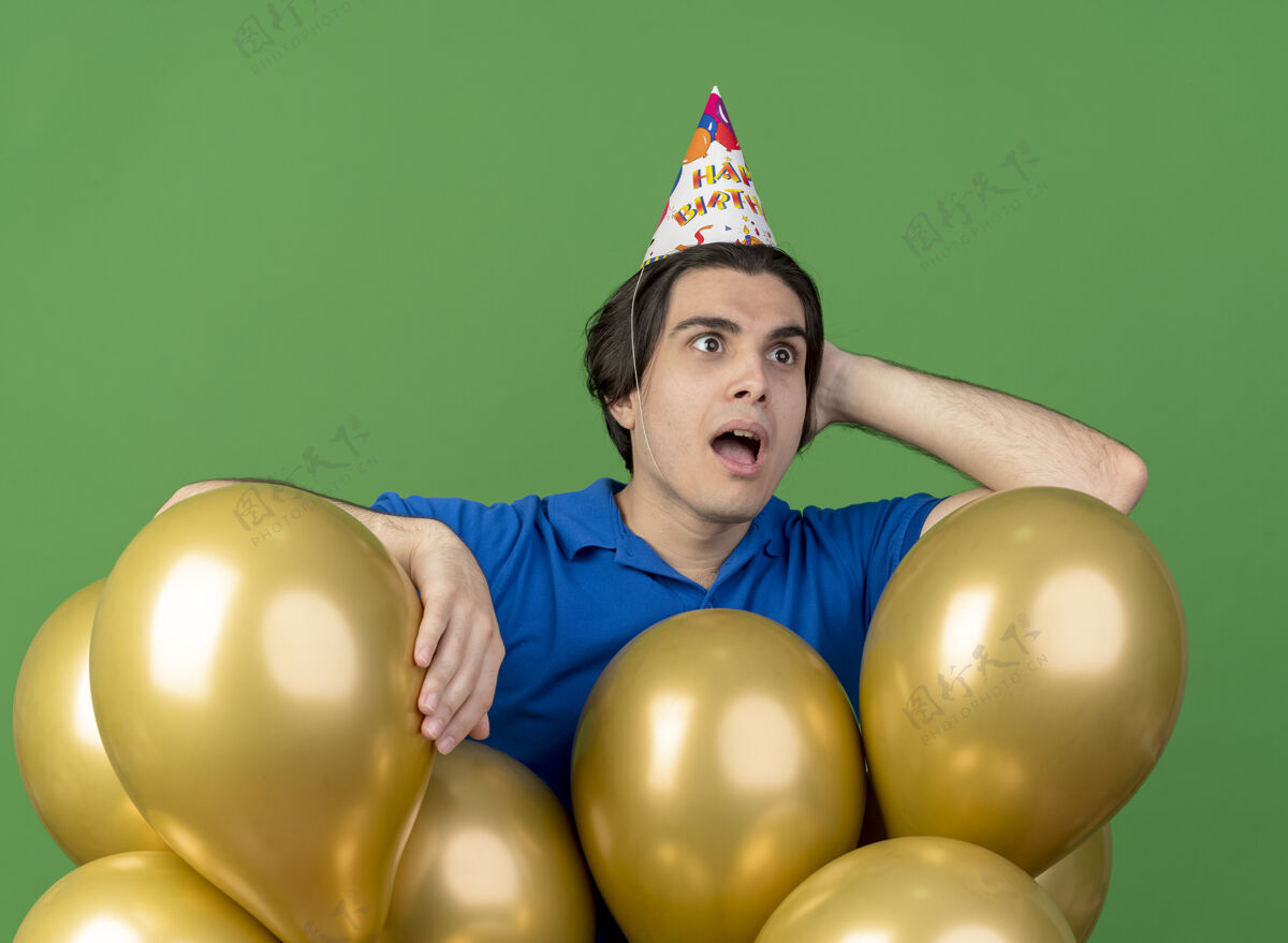 站戴着生日帽的白人帅哥惊讶地把手放在头上 后面站着氦气球惊喜氦穿