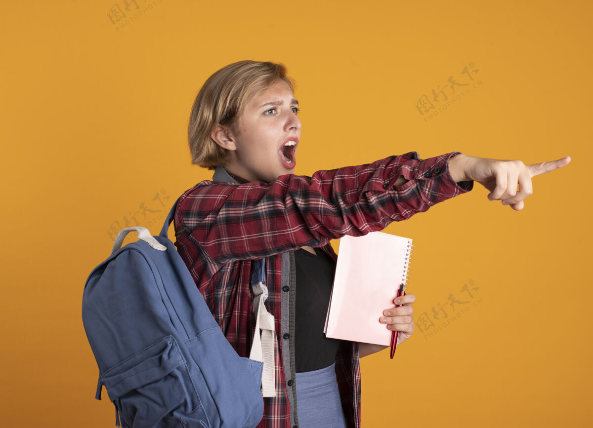 笔记本恼怒的年轻斯拉夫女学生戴着耳机 背着背包 拿着笔记本和笔 看着并指着旁边女孩学生年轻