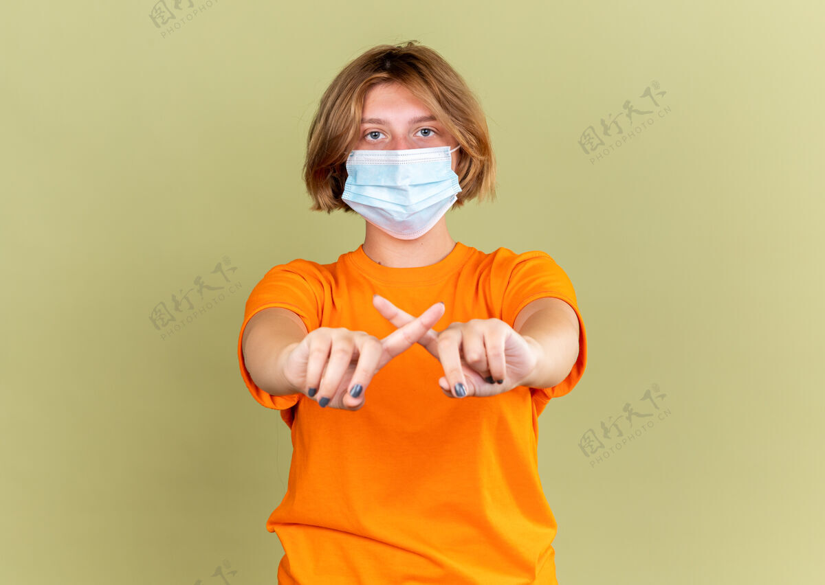手势不健康的年轻女子穿着橙色t恤 戴着护面面具 感觉不适 感染病毒 停止手势 食指交叉 站在绿色的墙上站立痛苦病毒