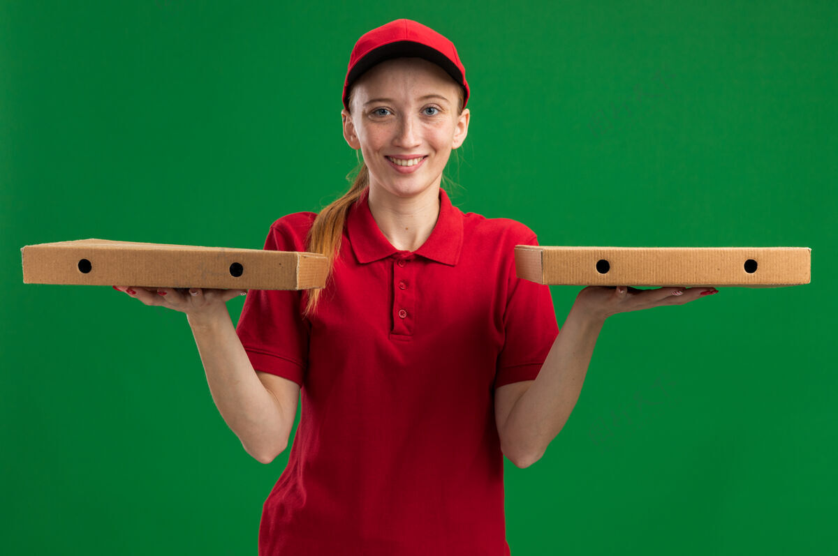 拿着身穿红色制服 头戴鸭舌帽 手持披萨盒的年轻送货女孩站在绿色的墙上 脸上带着幸福的微笑微笑年轻制服