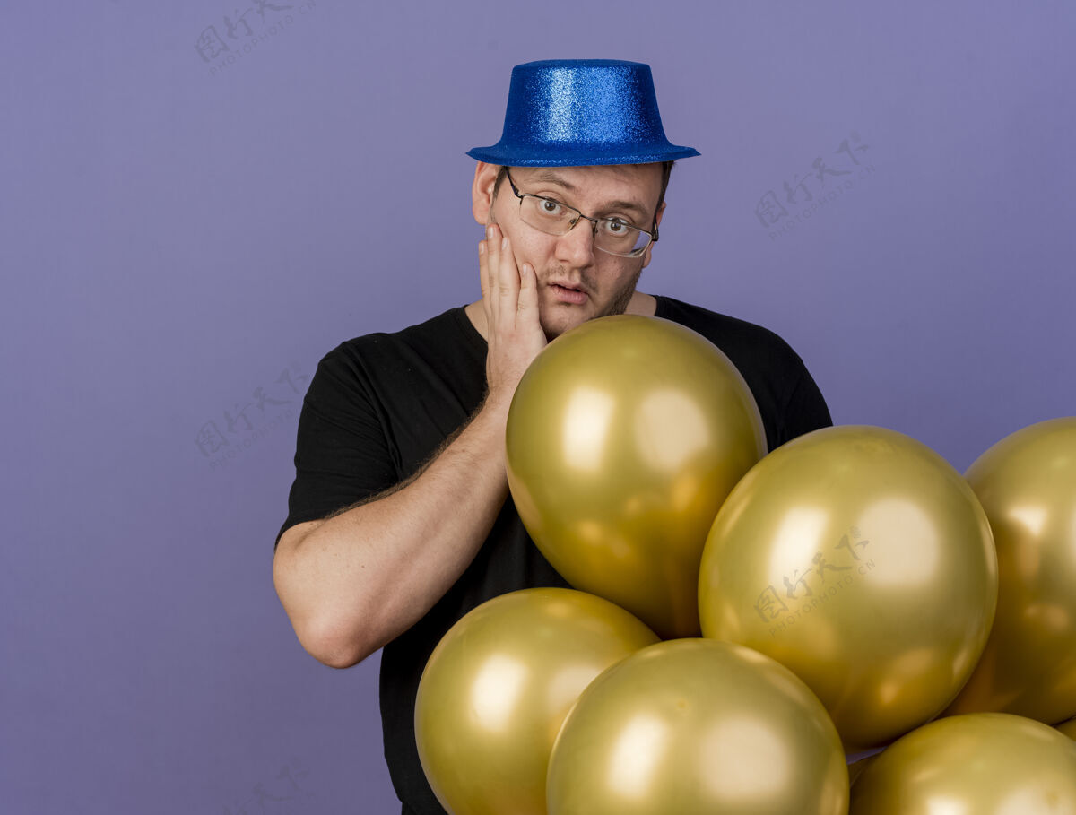 手令人印象深刻的成年斯拉夫人戴着眼镜 戴着蓝色派对帽 手放在脸上 站在那里放着氦气球光学眼镜脸
