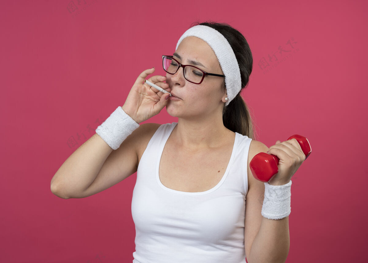 光学自信的年轻运动女孩戴着眼镜戴着头带和腕带拿着哑铃假装抽烟假装手持腕带
