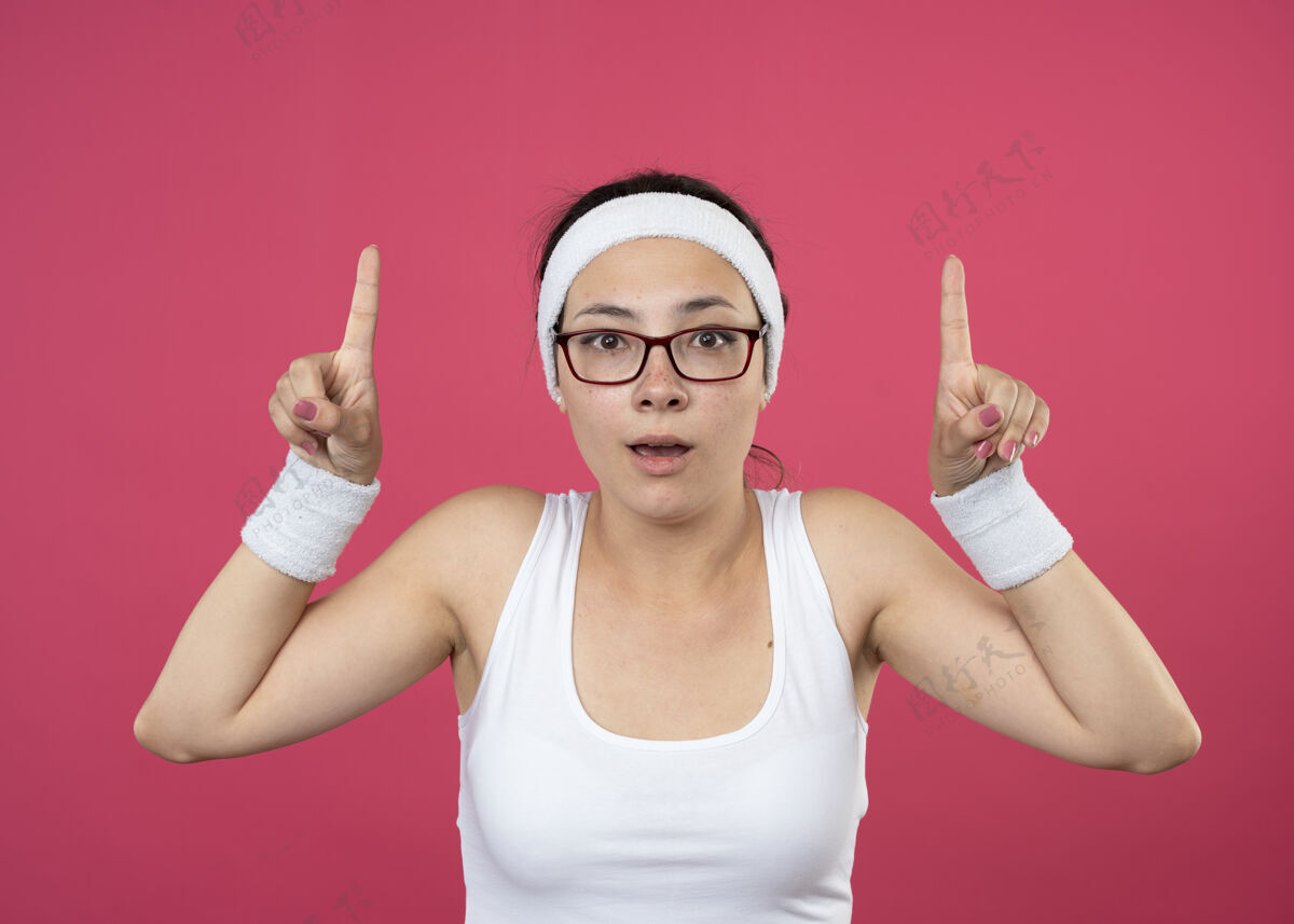 年轻惊讶的年轻运动女孩戴着眼镜戴着头带和腕带双手指向上穿二光学