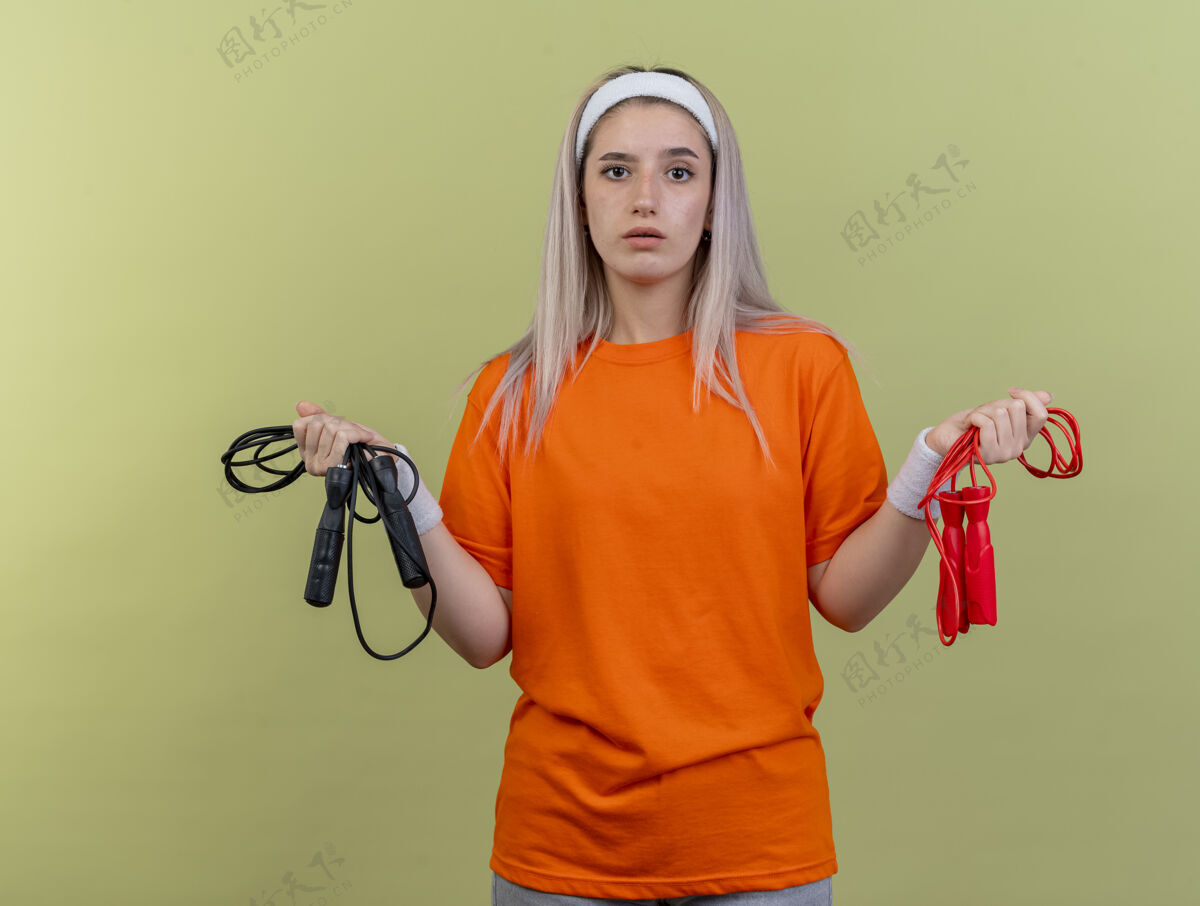 腕带困惑的年轻白人运动女孩戴着背带 戴着头带和腕带 手里拿着跳绳头带背带复制