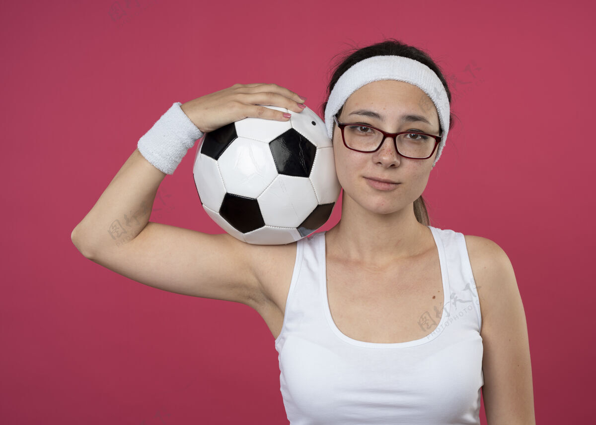 请戴着眼镜 戴着头带和腕带的快乐的年轻运动女孩把球放在肩上运动穿光学