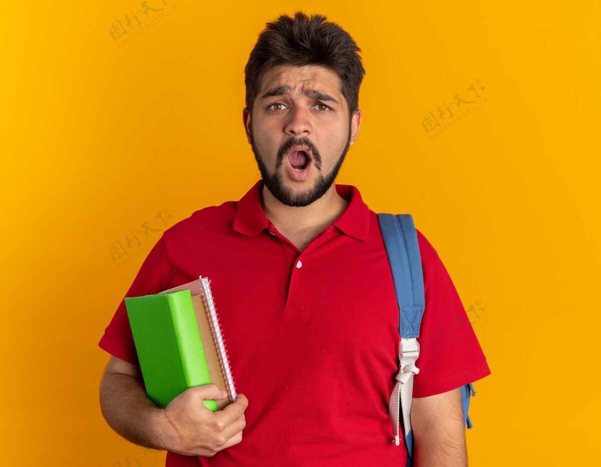 胡子年轻的留着胡子的学生 穿着红色马球衫 背着背包 拿着笔记本 站在那里显得困惑和惊讶小伙子站着迷茫