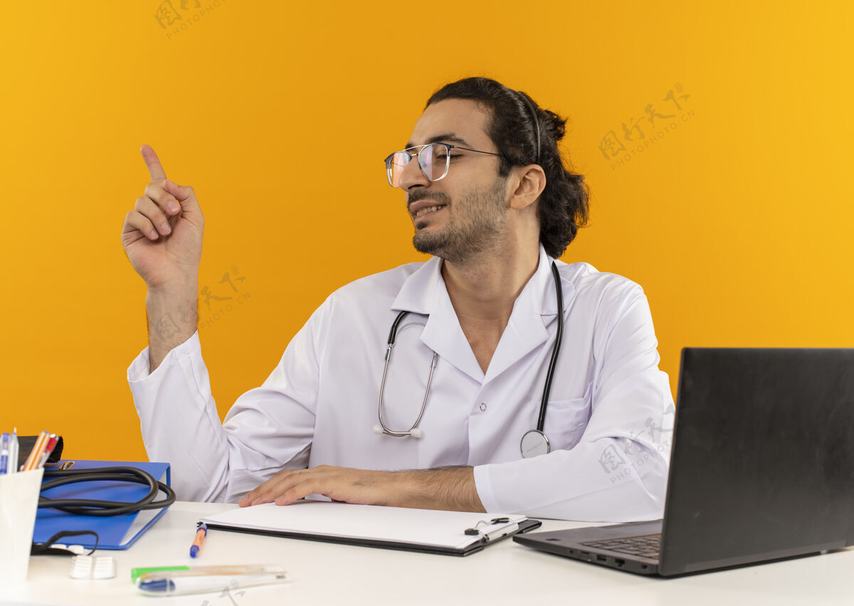 笔记本电脑面带微笑的年轻男医生戴着医用眼镜 穿着医用长袍 手持听诊器坐在办公桌旁工作眼镜穿