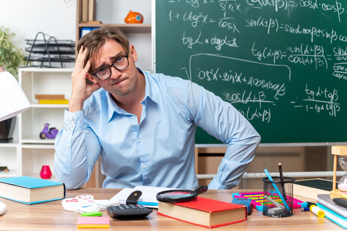 眼镜年轻的男老师戴着眼镜 表情混乱 坐在课桌旁 拿着书和笔记在教室的黑板前表情黑板前面
