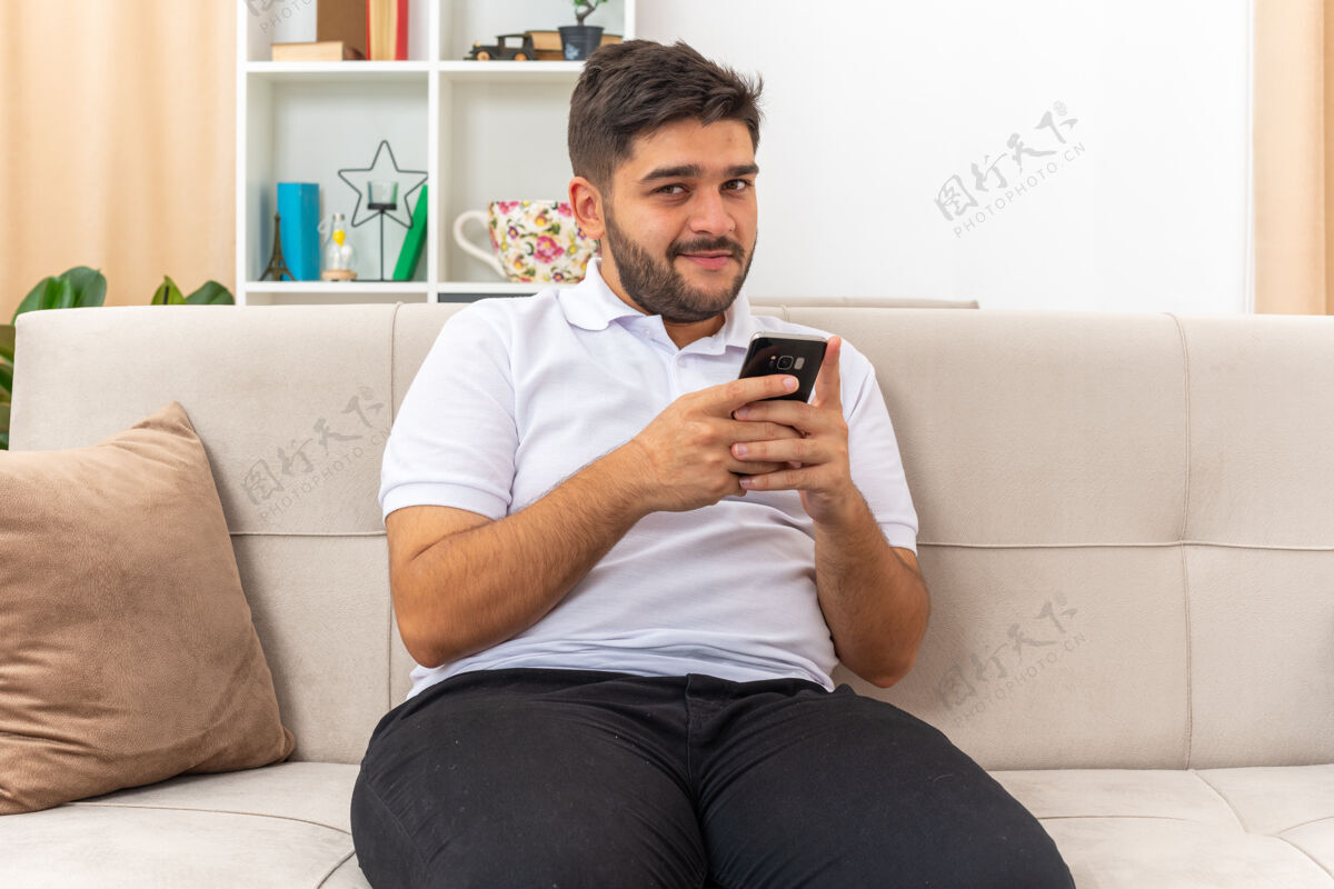 积极穿着休闲服的年轻人手里拿着智能手机 看上去很开心 脸上带着积极的笑容 自信地坐在客厅里的沙发上手机微笑坐着