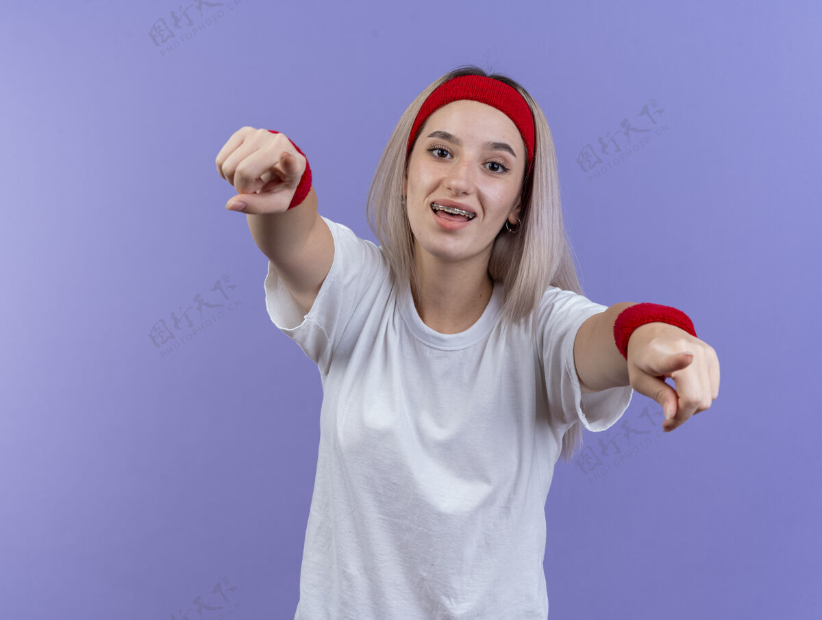 头带快乐的年轻白种人运动女孩戴着背带 戴着头带和腕带看着镜头 两只手放在紫色上尖腕带手
