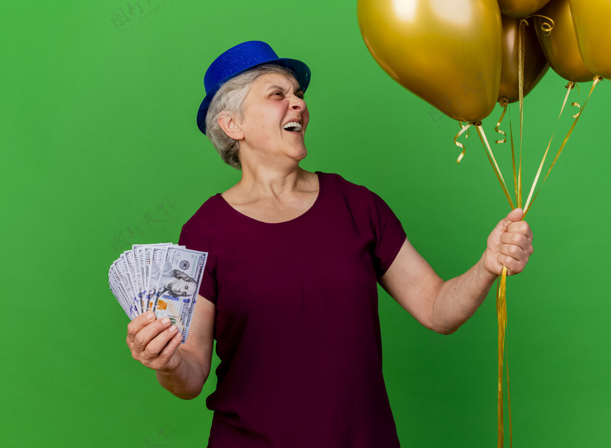钱戴着派对帽的老妇人手里拿着钱 看着绿色的氦气球气球欢笑老人