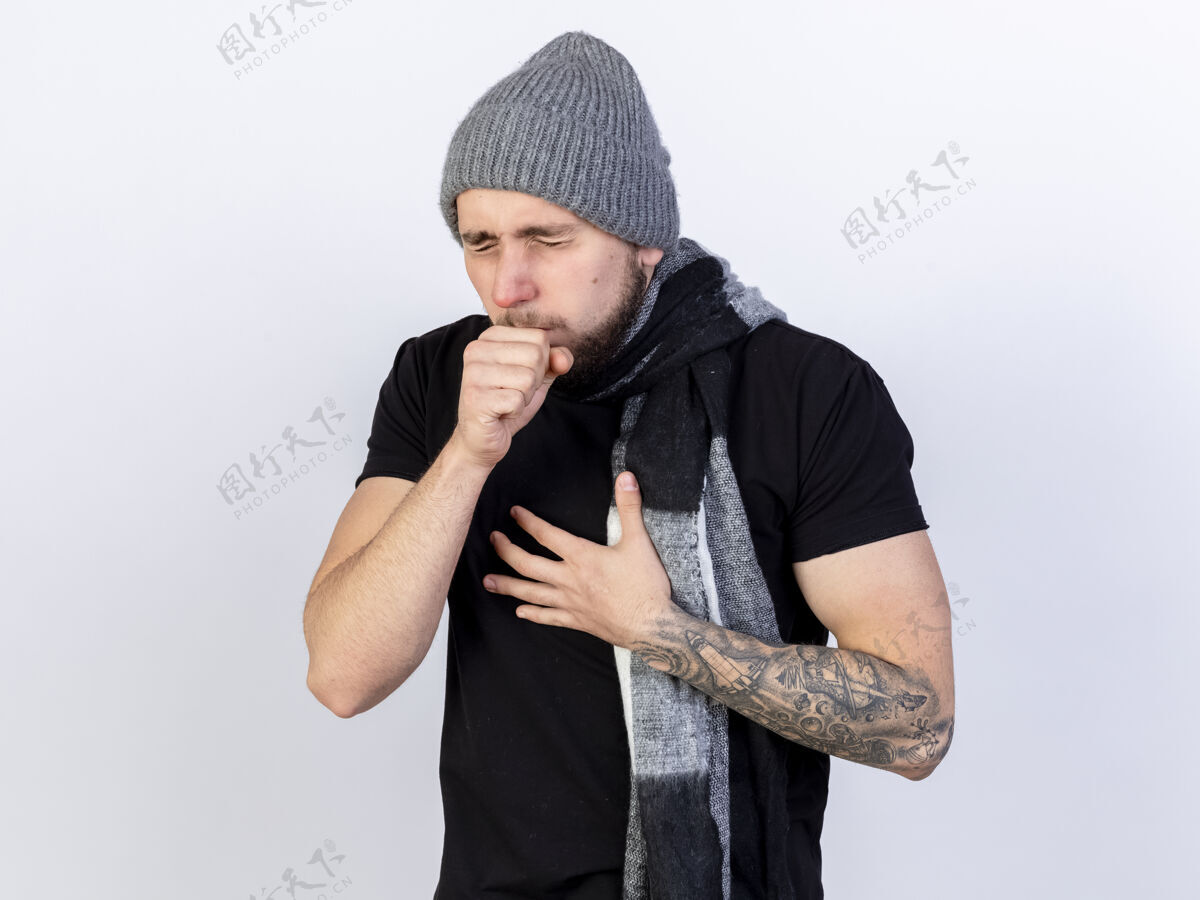 白色一个戴着冬日帽 戴着围巾 浑身酸痛的白人年轻人咳嗽着 把手放在白色的胸口上男人穿冬天