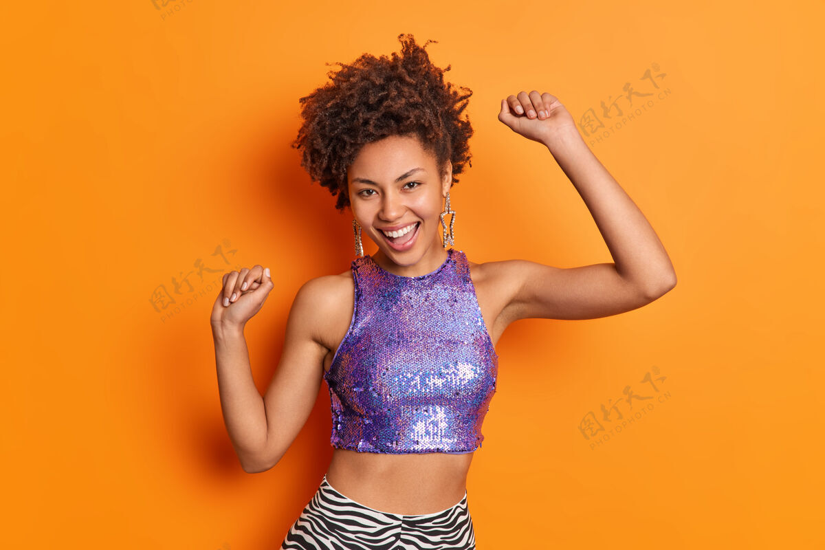 模特快乐时尚的美国黑人妇女卷发跳舞无忧无虑穿着紫色闪闪发光的上衣举起双臂孤立在生动的橙色墙壁乐趣头发快乐