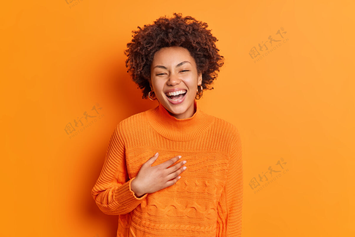 年轻真挚的情感和积极的情感概念快乐快乐的女人微笑着宽大的手放在胸前笑着有趣的故事穿着休闲毛衣隔离在橙色的墙上触摸快乐非洲