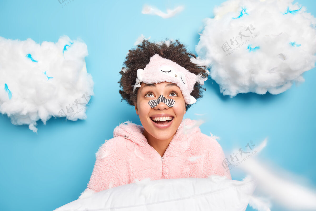早晨快乐的卷发年轻女子戴着口罩戴在鼻子上穿着睡衣高兴地看着上面微笑着宽大地抱着枕头准备睡觉 隔着蓝色的墙壁休息蒙眼柔软枕头