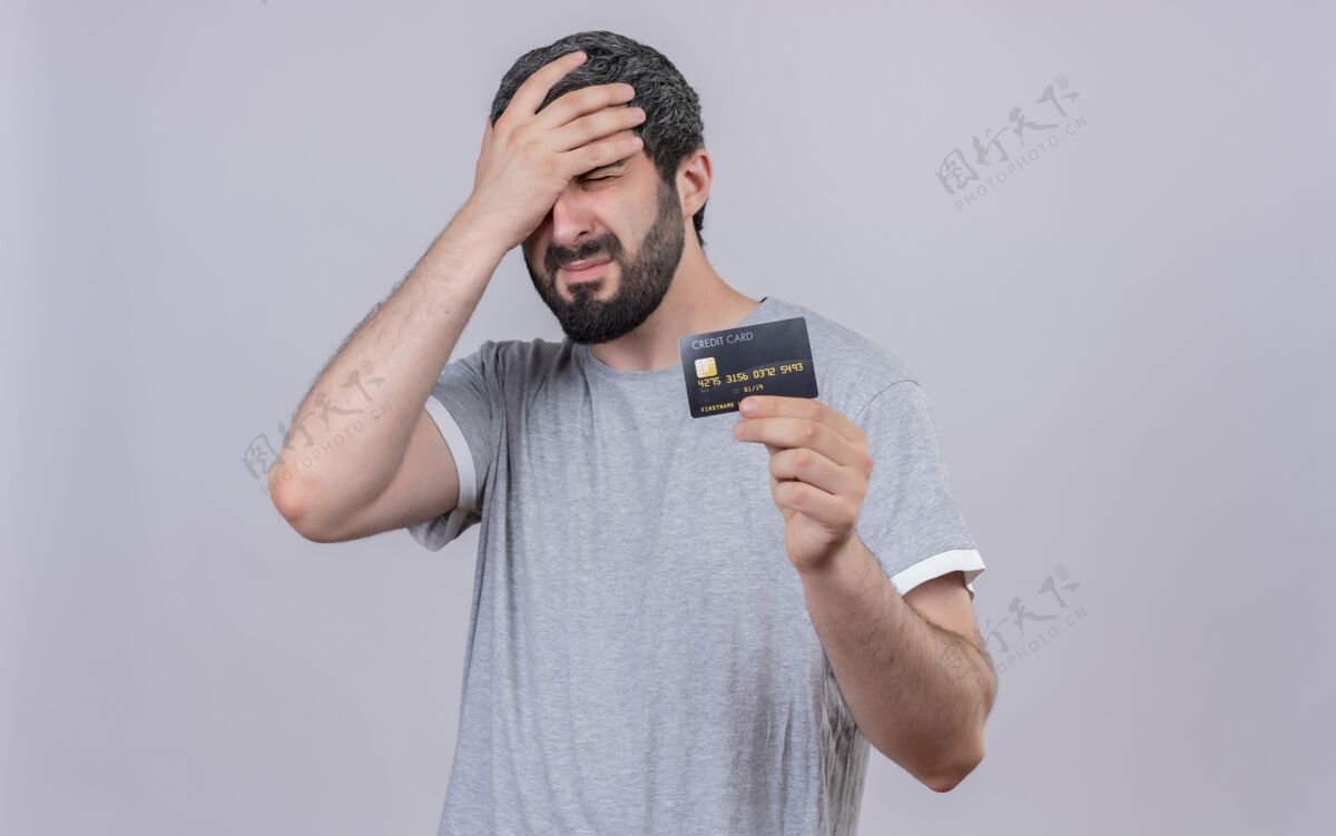 信用紧张的年轻帅哥拿着信用卡 把手放在头上闭着眼睛孤立在白墙上压力人手