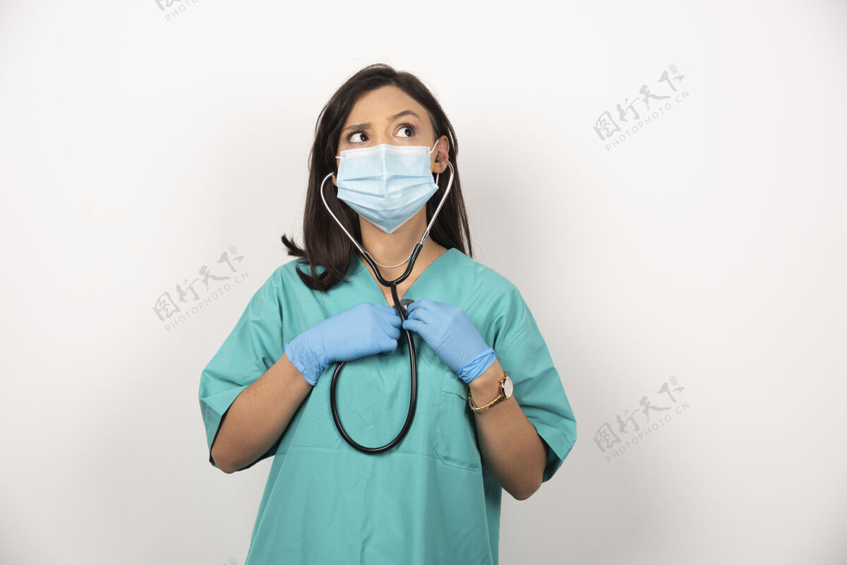 妇女年轻医生手持听诊器在白色背景上思考高品质照片站立医生面罩