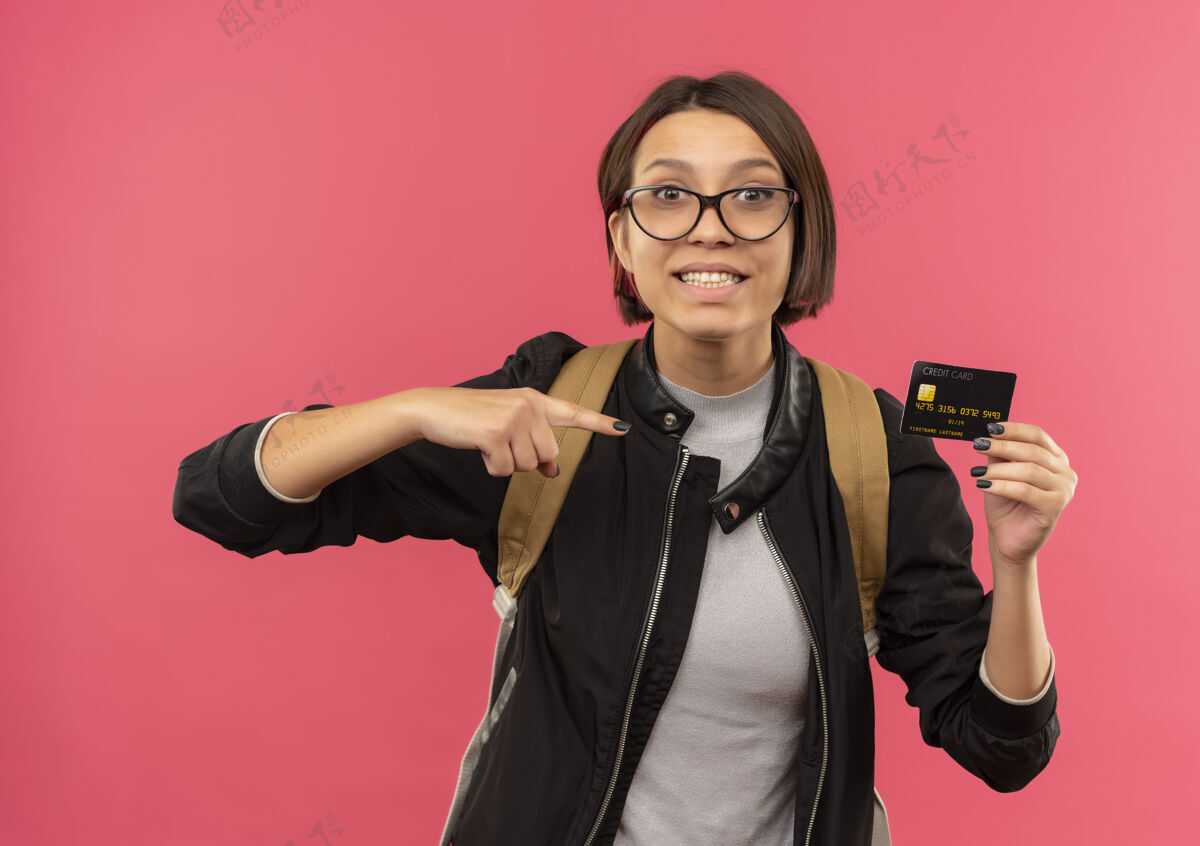 背面带微笑的年轻女学生戴着眼镜 背着书包 拿着信用卡指着隔离在粉红色墙上的墙信用人人