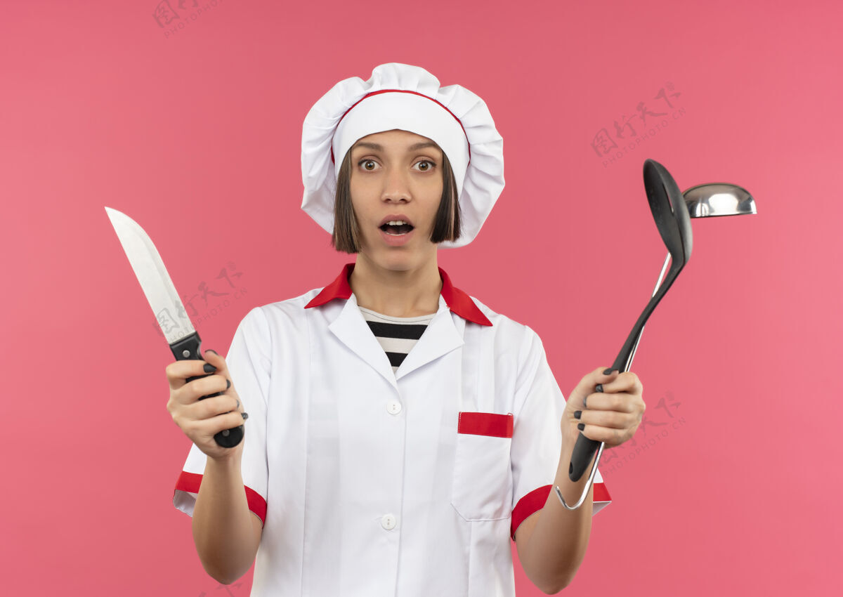 姿势惊讶的年轻女厨师穿着厨师制服拿着刀 抹刀和勺子隔离在粉红色的墙上女性烹饪惊喜