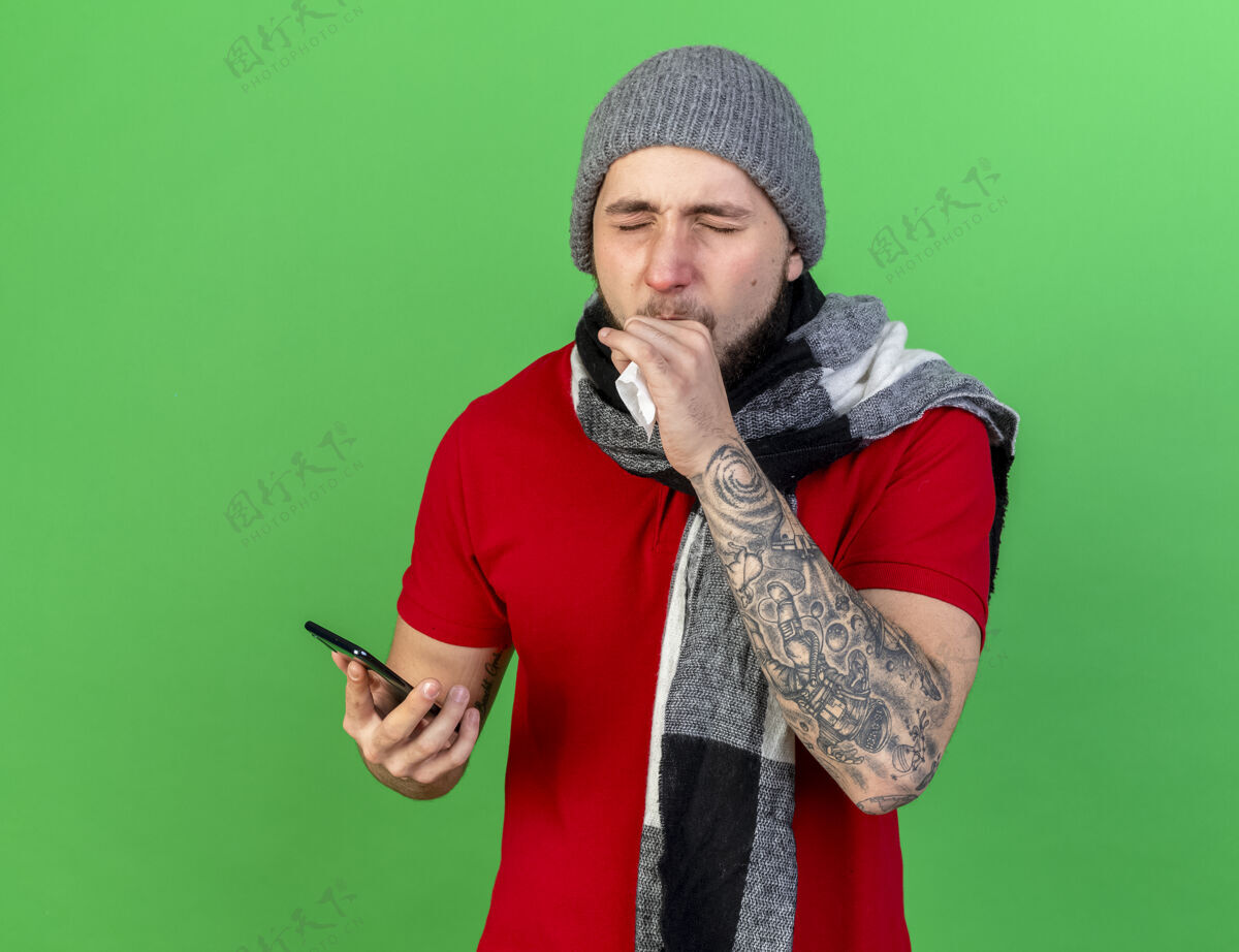 男人咳嗽的年轻白人病人戴着冬天的帽子和围巾拿着纸巾和电话帽子疾病围巾