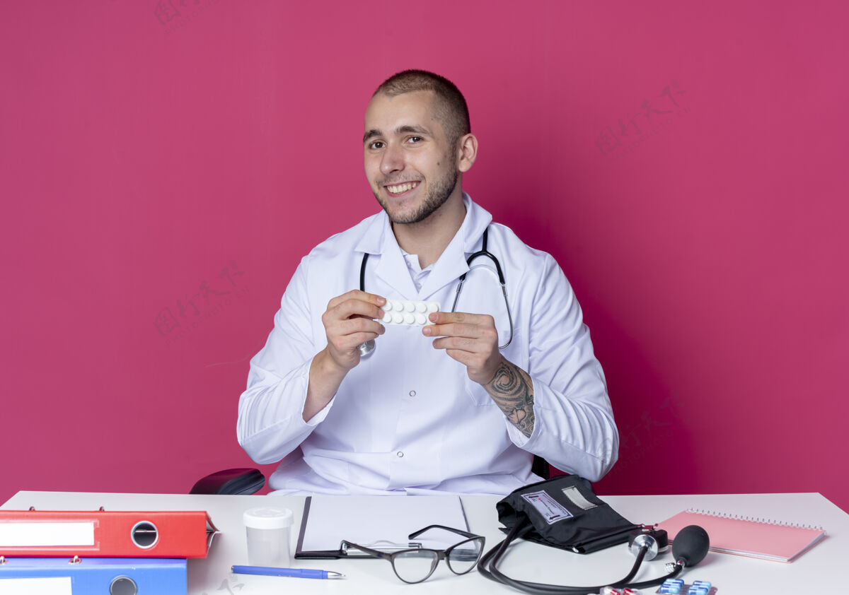 长袍面带微笑的年轻男医生 穿着医用长袍 听诊器 坐在办公桌旁 手里拿着工作工具 粉红色的墙上隔着一包药片年轻人人药片