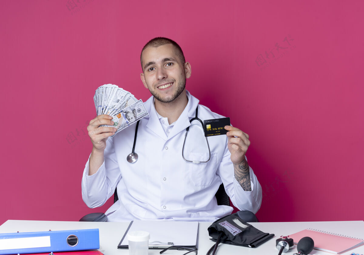 长袍面带微笑的年轻男医生穿着医用长袍 听诊器坐在办公桌旁 手里拿着工作工具 手里拿着信用卡和隔离在粉红色墙上的钱公民听诊器工作