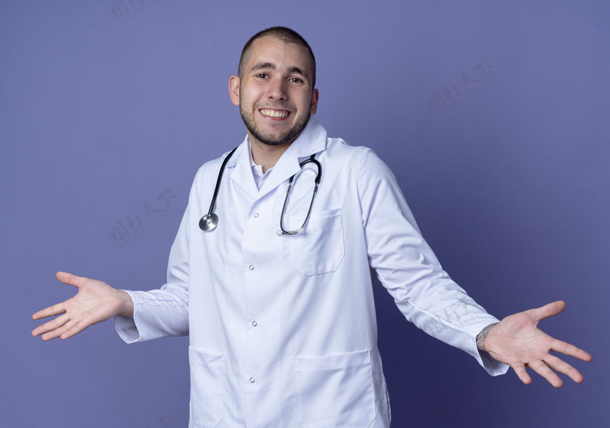 人面带微笑的年轻男医生穿着医用长袍 手持听诊器 双手空空地孤立在紫色的墙上年轻衣服公民
