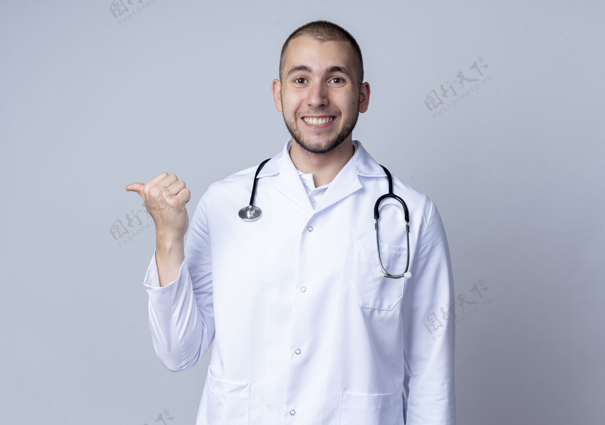 公民面带微笑的年轻男医生 身穿医用长袍 脖子上戴着听诊器 指着隔离在白墙上的一侧长袍医生穿着