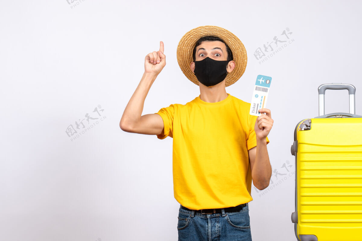 拿着正面图：戴草帽的年轻人站在黄色手提箱旁 拿着旅行票 手指向上男人视图站着