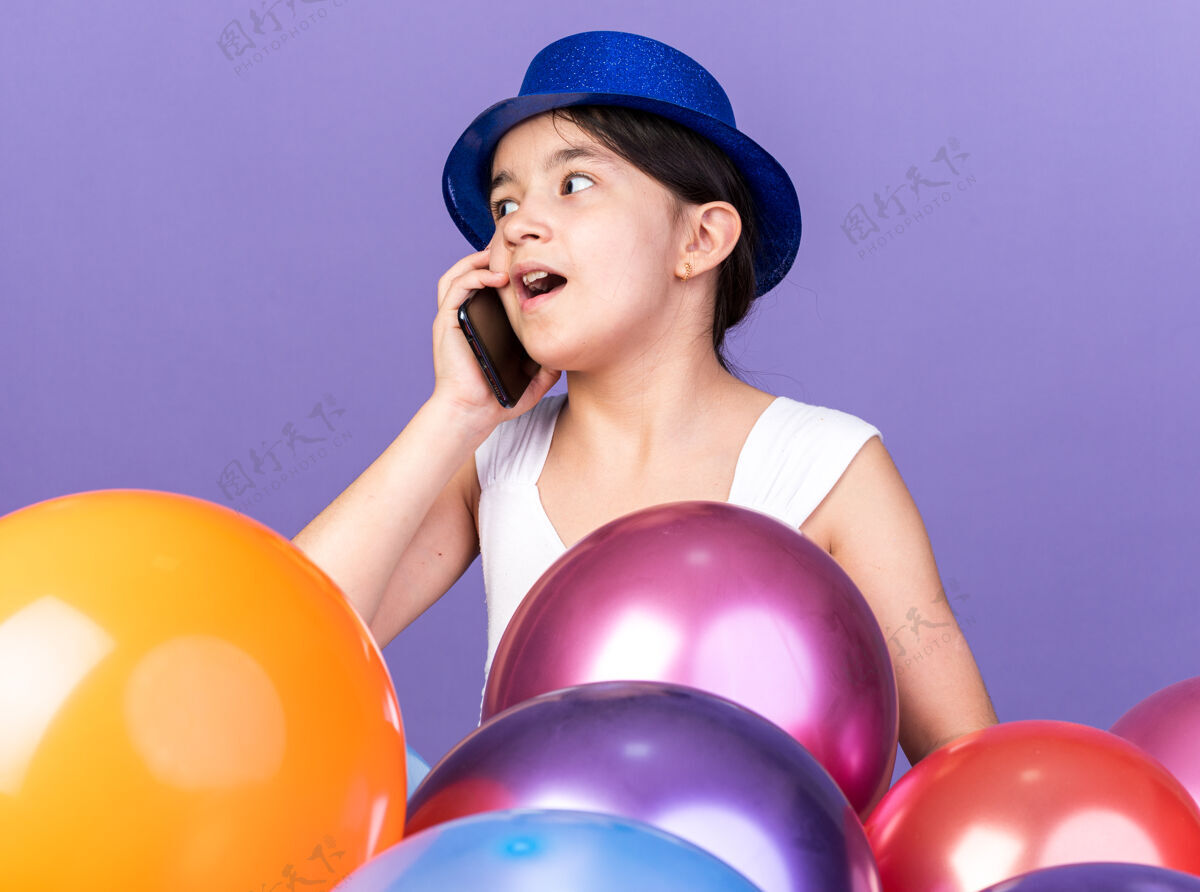 电话惊讶的年轻白人女孩戴着蓝色的派对帽 一边打电话一边看着站在紫色墙上的氦气球 还有复制空间氦壁板穿