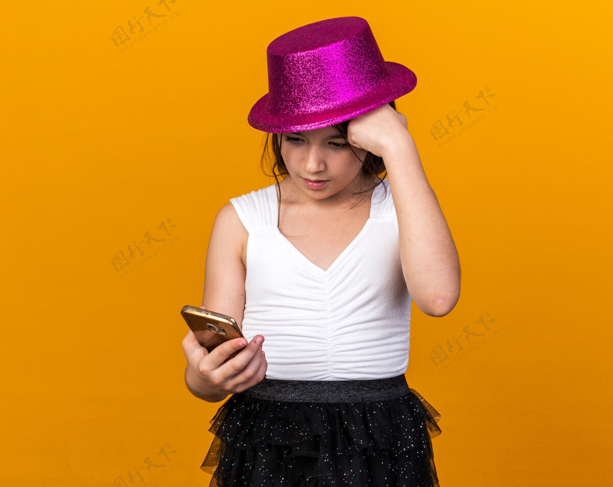 电话焦急的年轻白人女孩 戴着紫色的派对帽 把拳头放在额头上 拿着手机 看着隔离在橙色墙上的手机 还有复印空间年轻白种人拳头