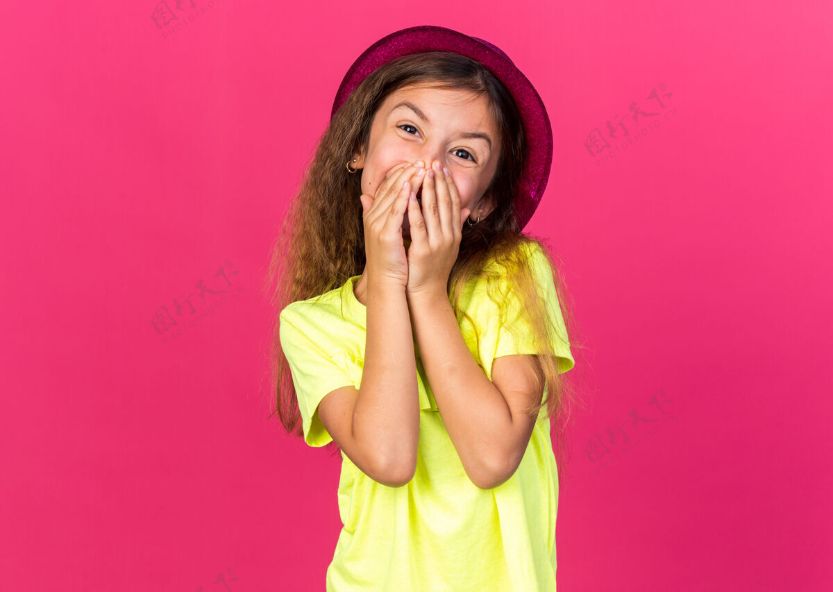小的可爱的白人小女孩 戴着紫色的派对帽 嘴巴被遮住 手被隔离在粉色的墙上 还有复印空间手帽子拜托