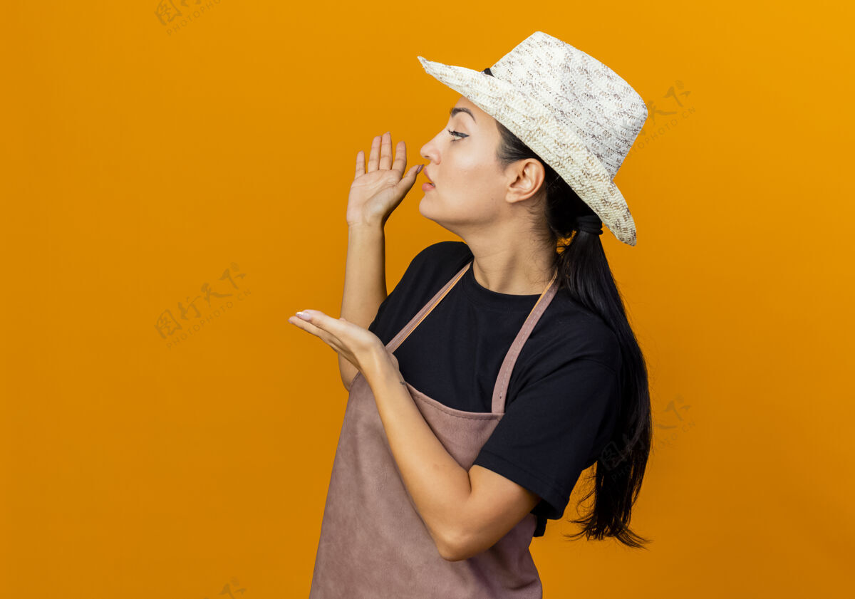 人穿着围裙 戴着帽子的年轻漂亮的女园丁站在橙色的墙上 双手放在身后展示着什么手脸花园