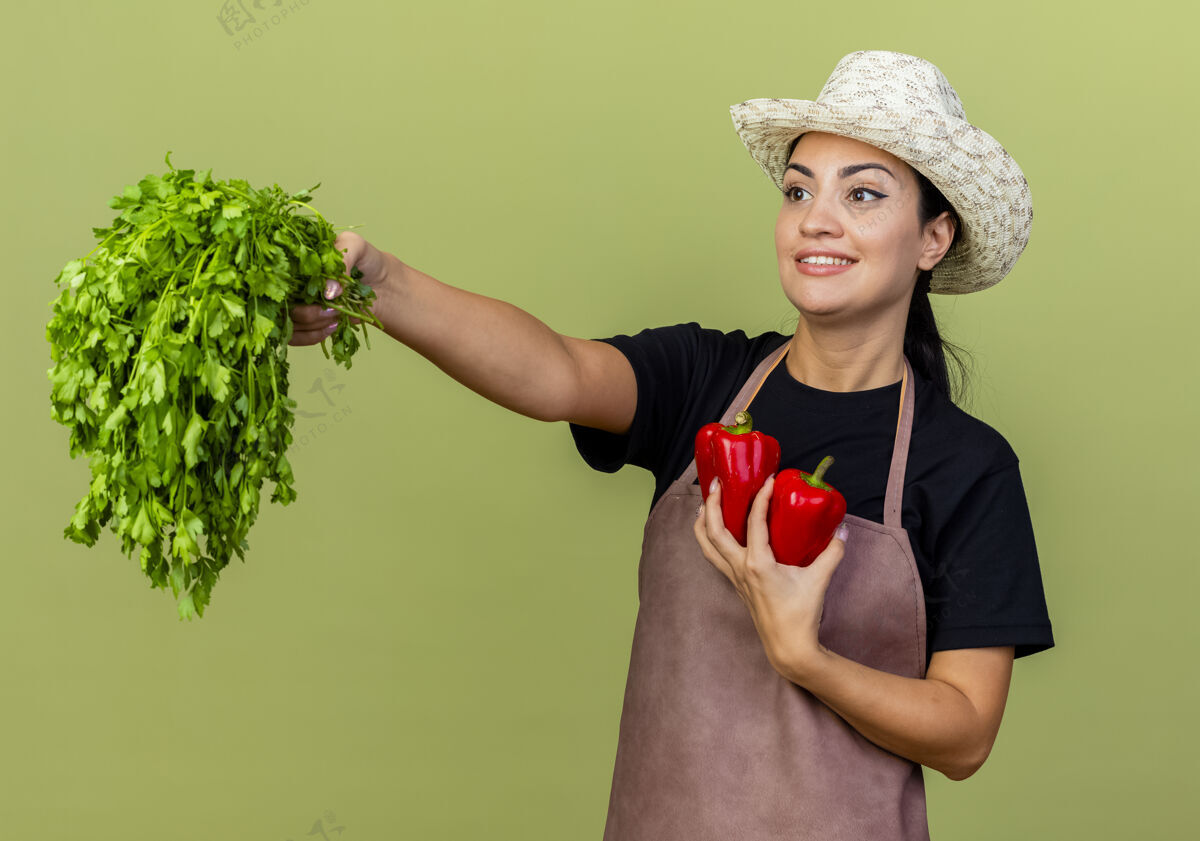 美丽年轻漂亮的女园丁 围着围裙 戴着帽子 手里拿着红甜椒和新鲜的草药 站在淡绿色的墙上欢快地笑着草药胡椒人