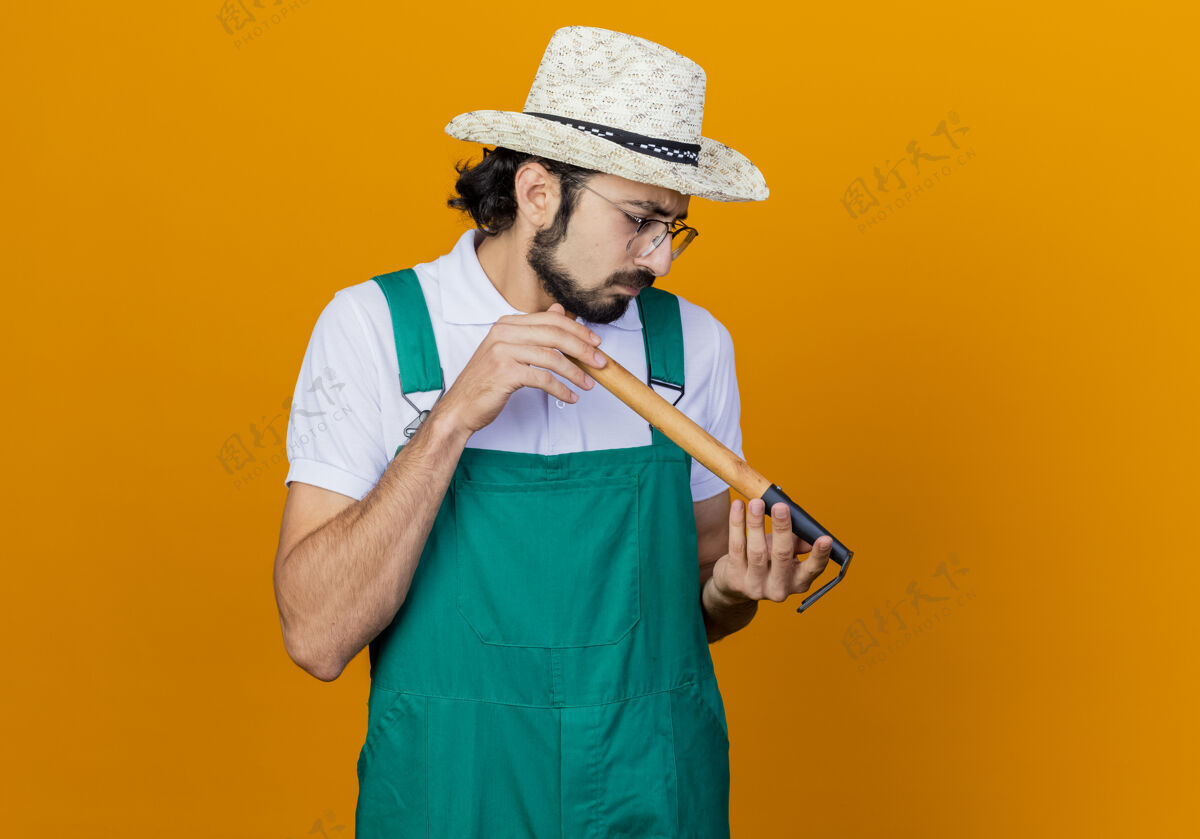 表情年轻的留着胡须的园丁 穿着连体衣 戴着帽子 手里拿着小耙子 站在橘色的墙上好奇地看着它人迷你抱着