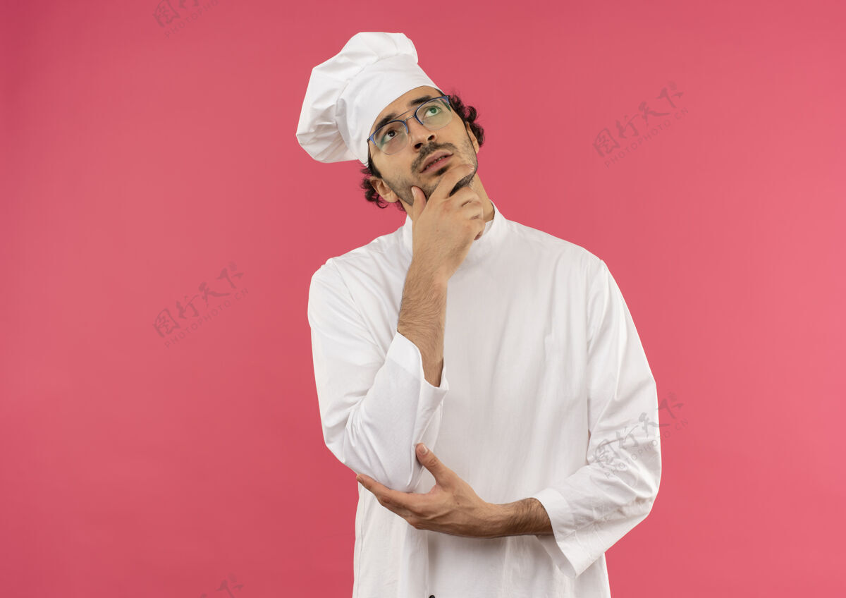 下巴看着一个年轻的男厨师 穿着厨师制服 戴着眼镜 手放在下巴上背景制服眼镜