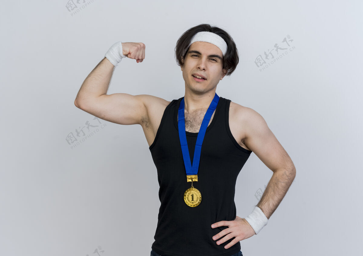 信心年轻的运动型男子身穿运动服 头戴金牌 脖子上举着拳头 站在白色的墙上 二头肌看起来很自信脸姿势提高