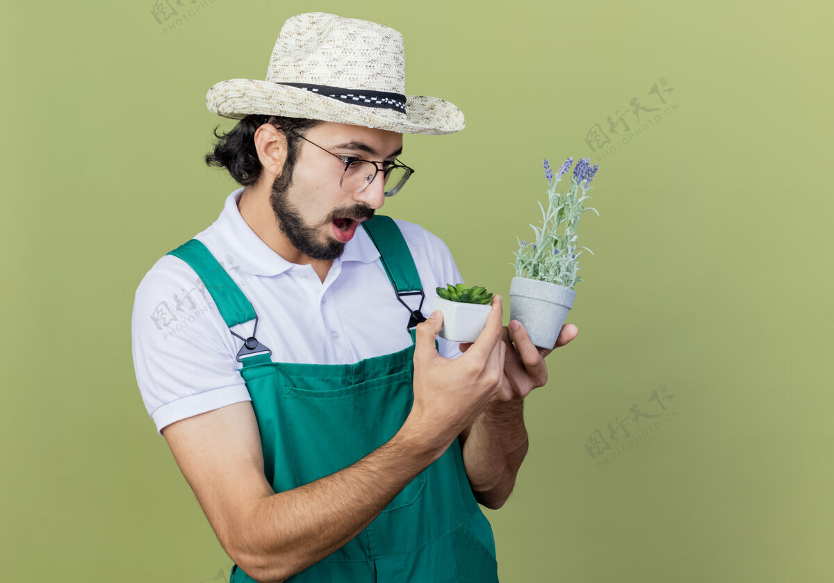 连身衣年轻的留着胡须的园丁 穿着连体衣 戴着帽子 手里拿着盆栽植物 站在浅绿色的墙上 看着它们惊讶不已胡子站着脸