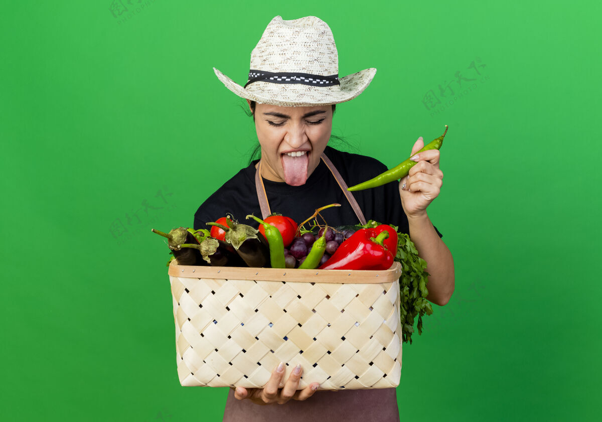 脸年轻漂亮的女园丁 围着围裙 戴着帽子 手里拿着装满蔬菜和青椒的篮子 站在绿色的墙上 带着厌恶的表情看着它感觉年轻美丽