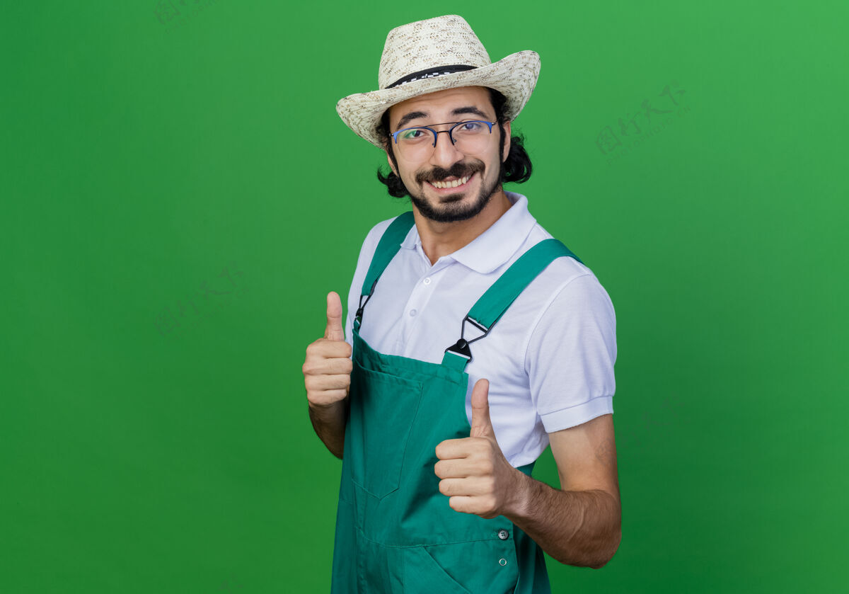 脸年轻的留着胡须的园丁 穿着连体衣 戴着帽子 面带微笑 站在绿色的墙上竖起大拇指连身衣帽子感觉
