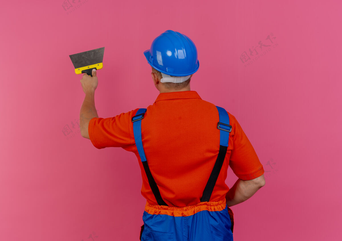 制服站在后面看年轻的男建筑工人穿着制服和安全帽手持油灰刀视图手持头盔