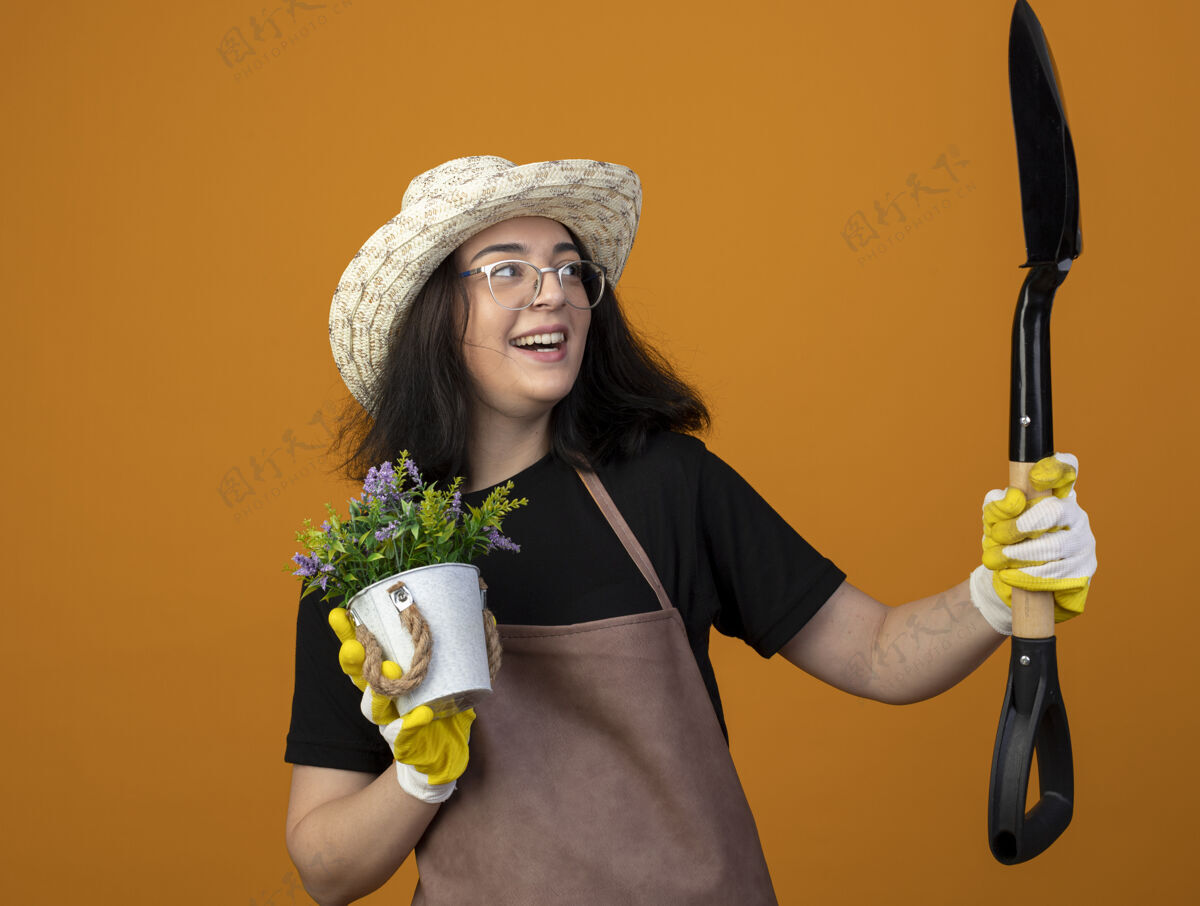 眼镜戴着眼镜 穿着制服 戴着园艺帽和手套的快乐的年轻黑发女园丁手持花盆 看着隔离在橙色墙上的铲子眼镜园艺人