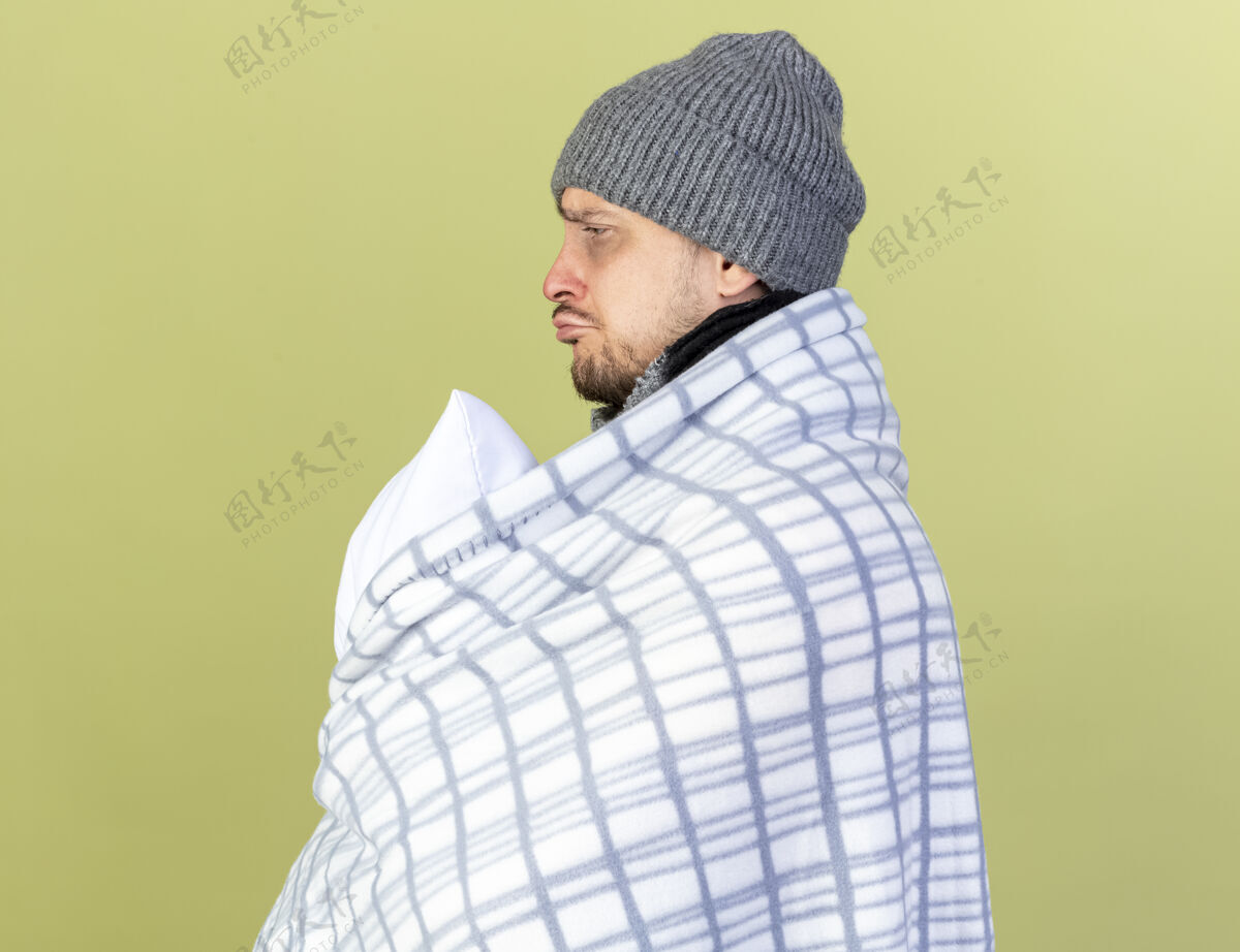 摆姿势悲伤的年轻金发病男子戴着冬天的帽子和围巾包裹在格子布站在一旁举行橄榄绿墙上孤立的枕头站着橄榄年轻