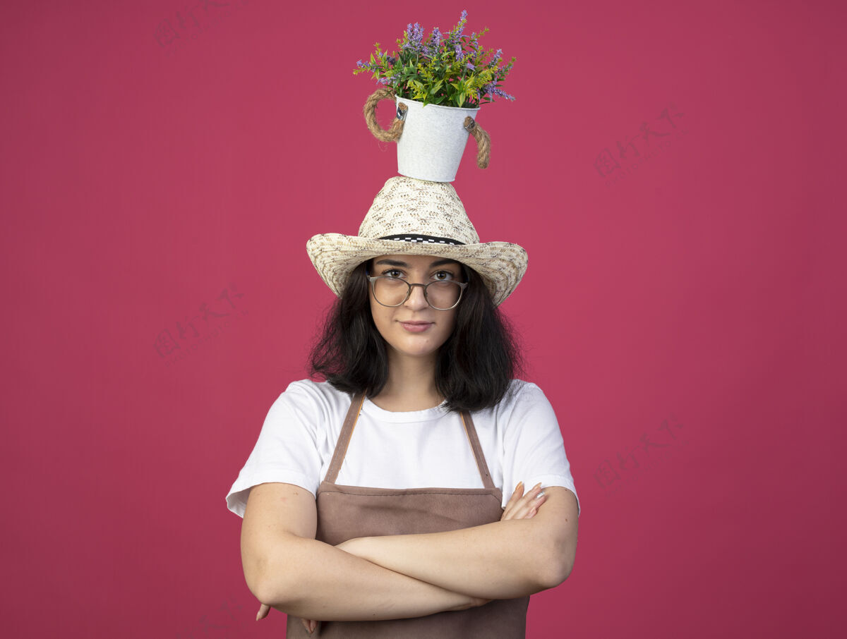 眼镜自信的年轻黑发女园丁 戴着眼镜 穿着制服 戴着园艺帽 双臂交叉 头上抱着花盆 隔离在粉红色的墙上光学自信人