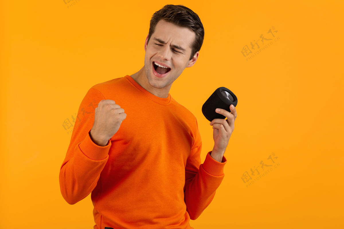 肖像穿着橙色毛衣的时髦微笑的年轻人拿着无线扬声器快乐地听音乐享受橙色的乐趣城市乐趣姿势
