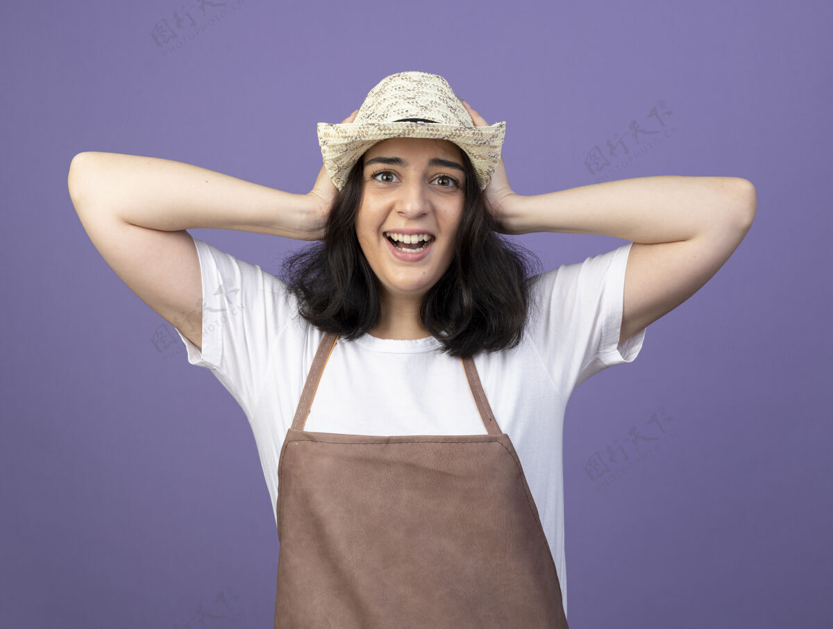 人兴奋的年轻黑发女园丁穿着制服戴着园艺帽把手放在紫色墙上孤立的头上市民女性人