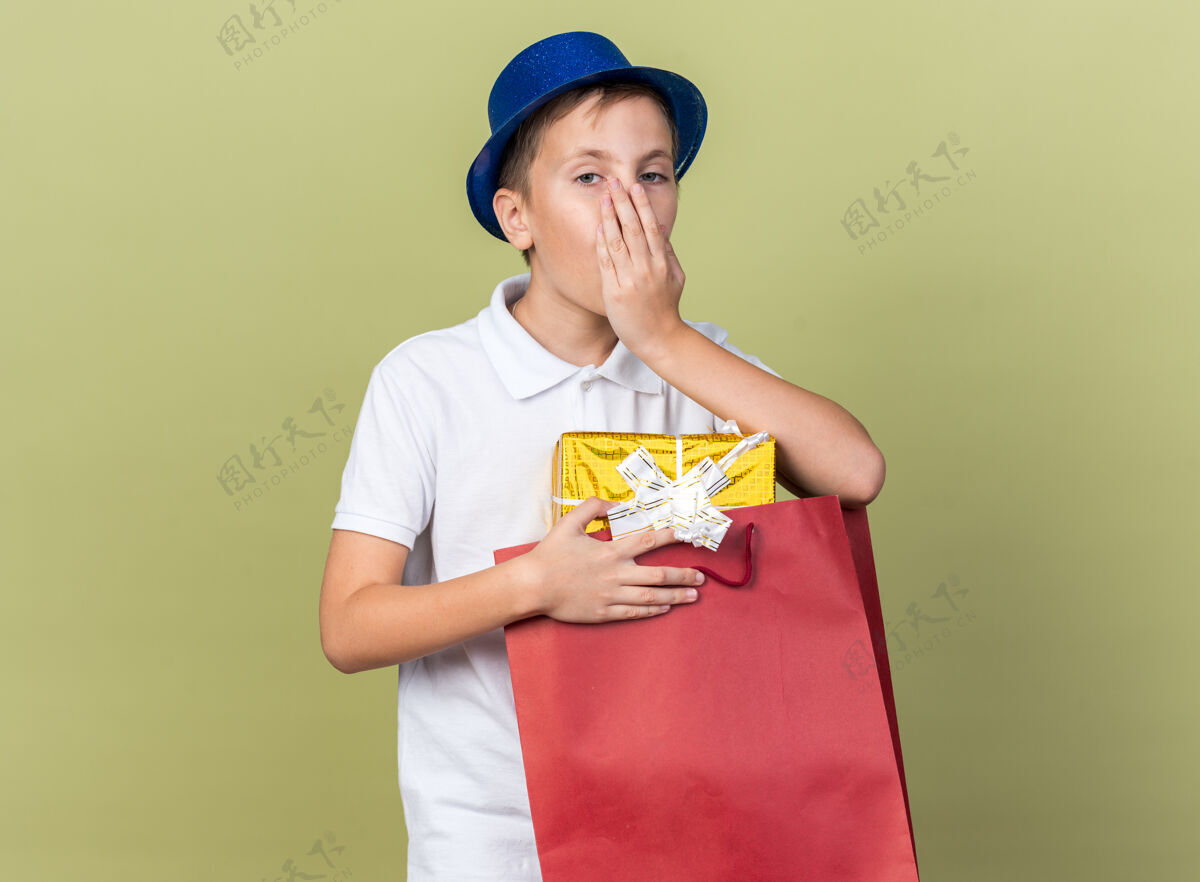 男孩惊讶的年轻斯拉夫男孩 戴着蓝色派对帽 手放在嘴上 拿着购物袋中的礼品盒 隔离在橄榄绿的墙上 留有复印空间包帽子惊喜