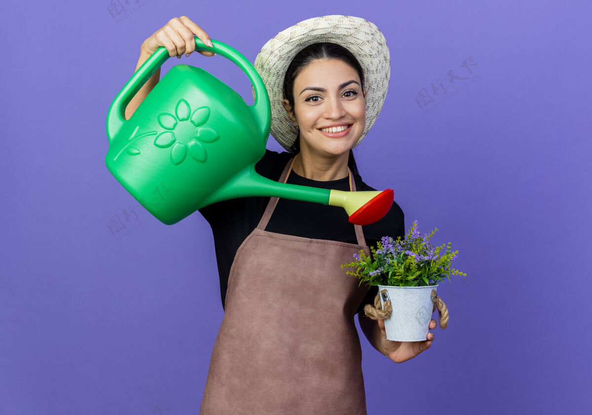 人年轻漂亮的女园丁围着围裙 戴着帽子 手里拿着浇水罐 站在蓝色的墙上 满脸笑容地浇灌着盆栽植物姿势漂亮人