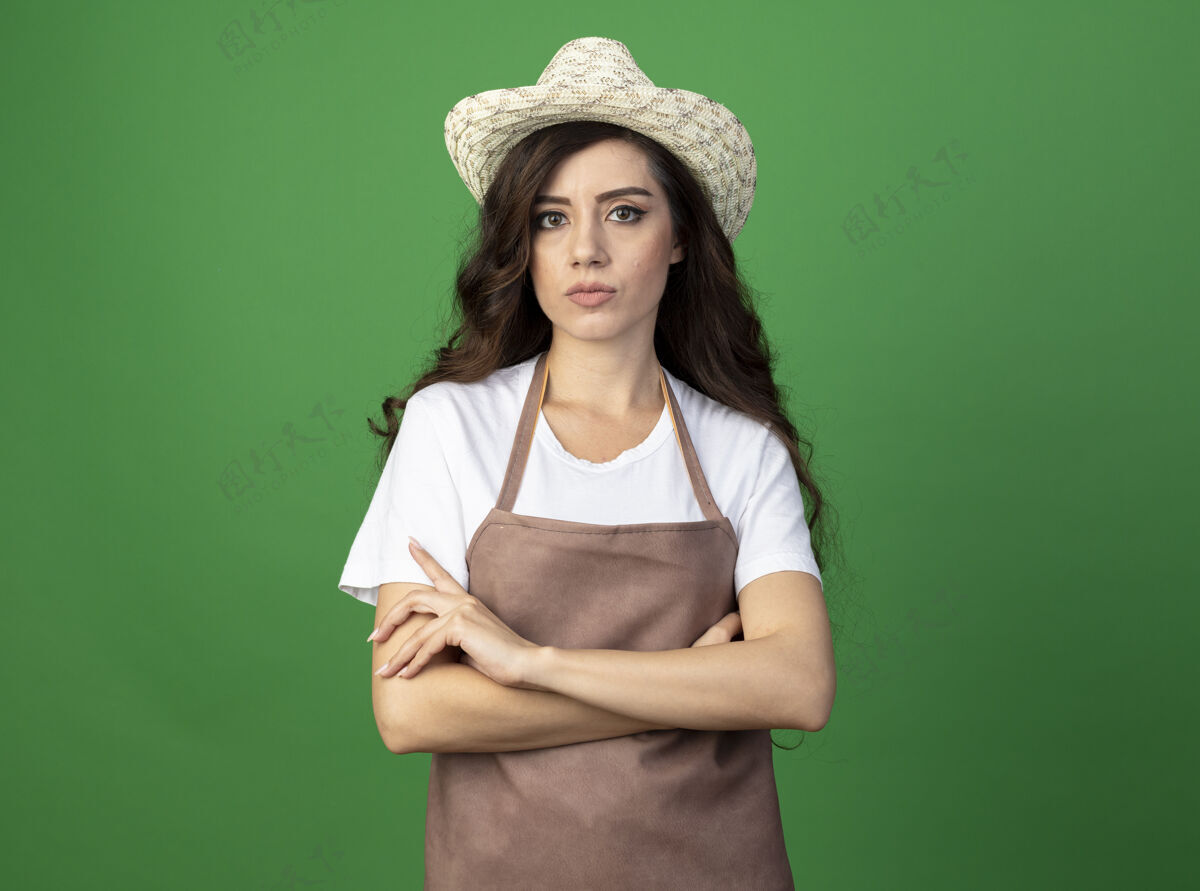 年轻人自信的年轻女园丁身着制服 戴着园艺帽 双臂交叉 孤零零地站在绿色的墙上姿势花园制服